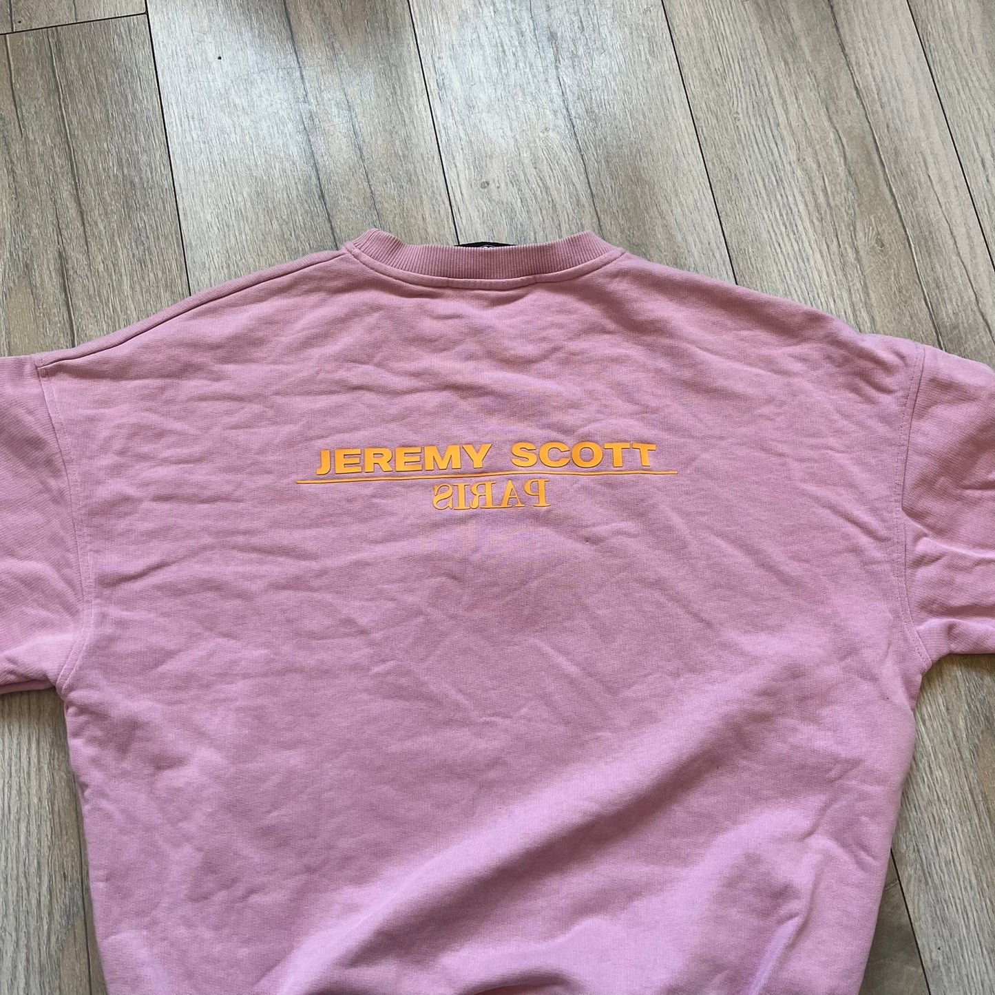 Jeremy Scott 2018 Popples Pink Crewneck is a pink sweatshirt featuring a front graphic of an upside-down orange Popples character with green hair and blue/red heart-shaped ears, finished with a glittery, shiny application. The upper back includes a rubberized orange “Jeremy Scott Paris” logo.
