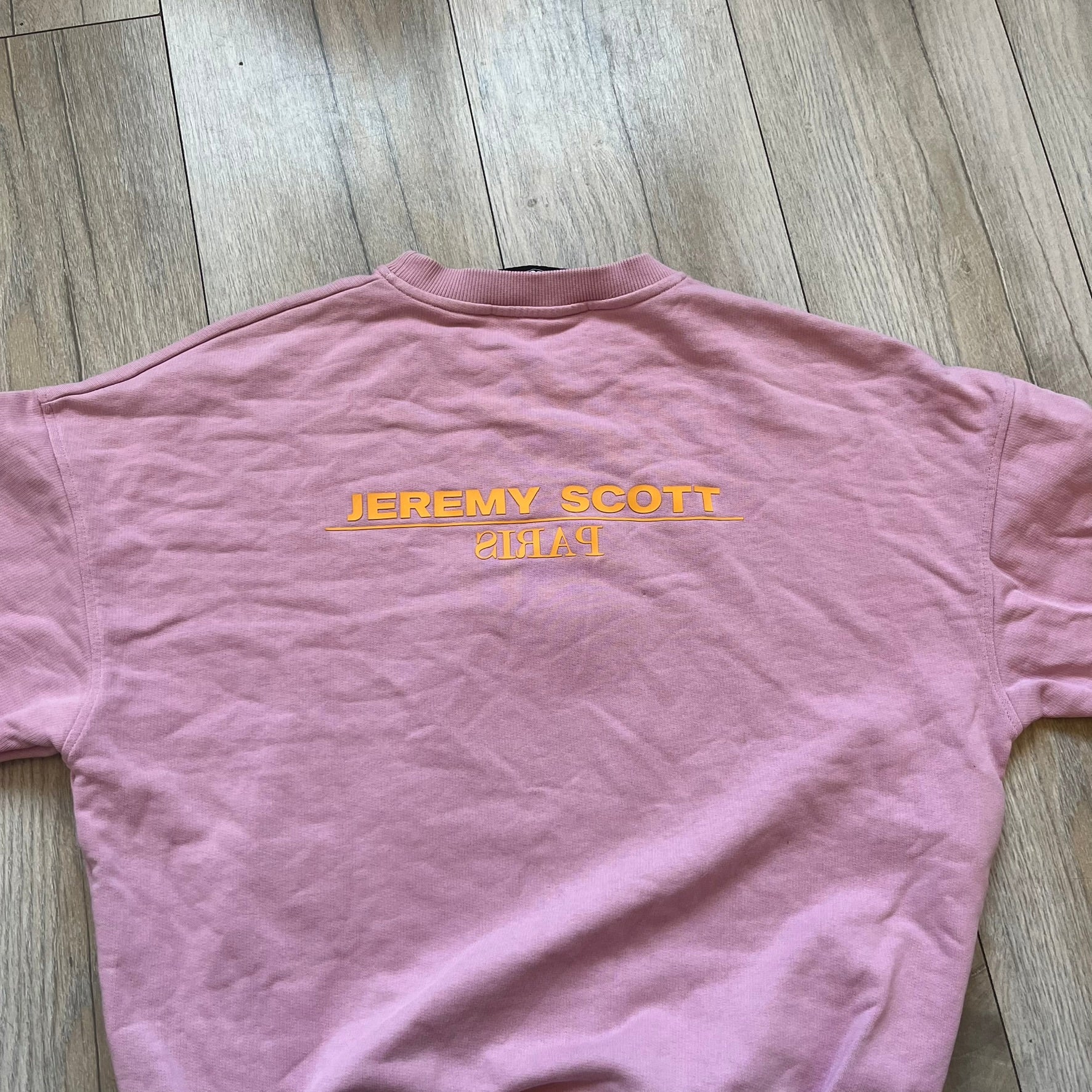 Jeremy Scott 2018 Popples Pink Crewneck is a pink sweatshirt featuring a front graphic of an upside-down orange Popples character with green hair and blue/red heart-shaped ears, finished with a glittery, shiny application. The upper back includes a rubberized orange “Jeremy Scott Paris” logo.
