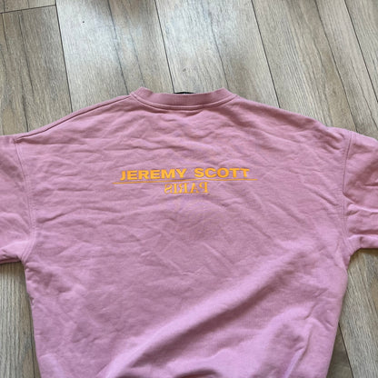 Jeremy Scott 2018 Popples Pink Crewneck is a pink sweatshirt featuring a front graphic of an upside-down orange Popples character with green hair and blue/red heart-shaped ears, finished with a glittery, shiny application. The upper back includes a rubberized orange “Jeremy Scott Paris” logo.
