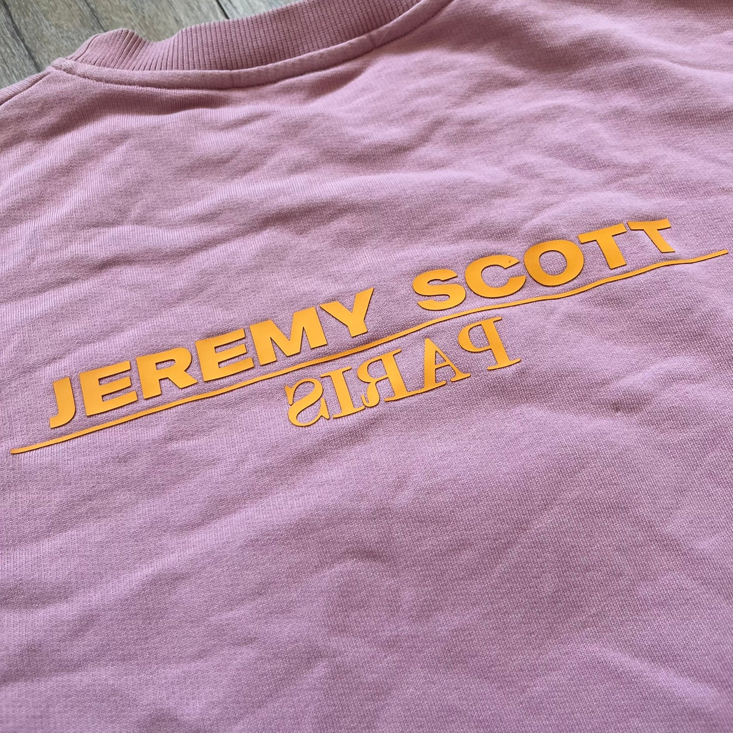 Jeremy Scott 2018 Popples Pink Crewneck is a pink sweatshirt featuring a front graphic of an upside-down orange Popples character with green hair and blue/red heart-shaped ears, finished with a glittery, shiny application. The upper back includes a rubberized orange “Jeremy Scott Paris” logo.
