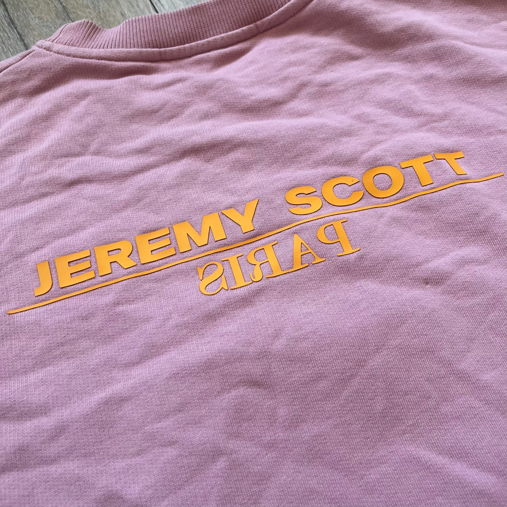 Jeremy Scott 2018 Popples Pink Crewneck is a pink sweatshirt featuring a front graphic of an upside-down orange Popples character with green hair and blue/red heart-shaped ears, finished with a glittery, shiny application. The upper back includes a rubberized orange “Jeremy Scott Paris” logo.
