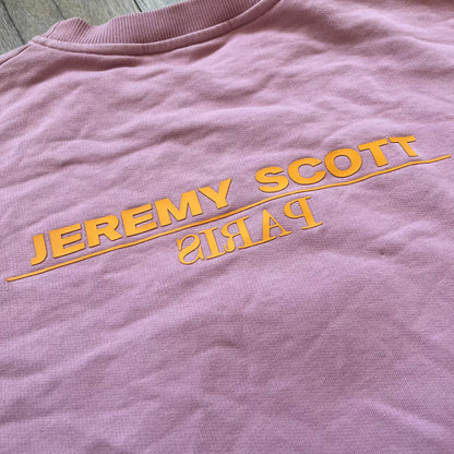 Jeremy Scott 2018 Popples Pink Crewneck is a pink sweatshirt featuring a front graphic of an upside-down orange Popples character with green hair and blue/red heart-shaped ears, finished with a glittery, shiny application. The upper back includes a rubberized orange “Jeremy Scott Paris” logo.
