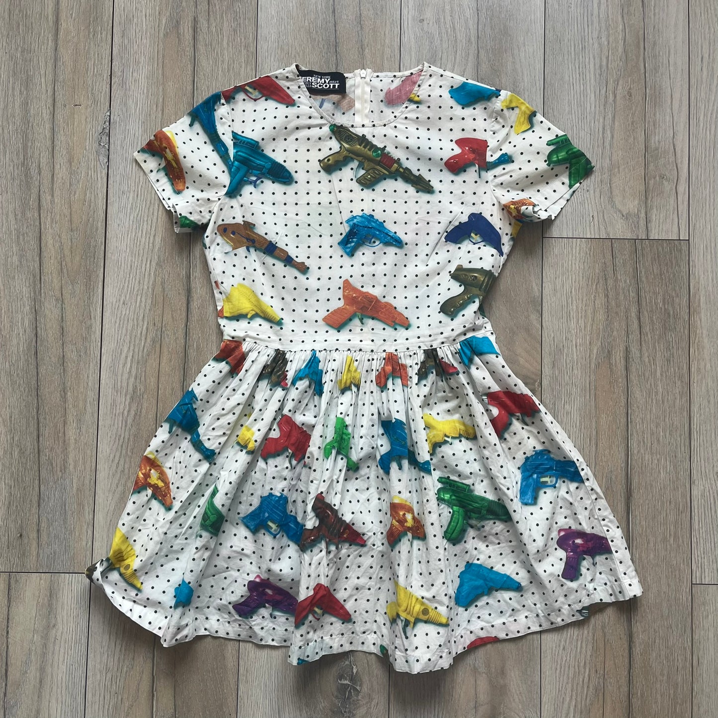 Jeremy Scott SS2016 Watergun Dress is a white flared short dress featuring an all-over print of multicolored realistic waterguns in varying sizes and orientations. The design is set against a black dotted pattern layered across the fabric. It has a fitted upper section with a flared skirt silhouette and includes a black Jeremy Scott neck tag.