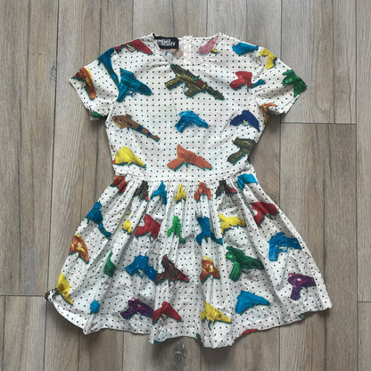 Jeremy Scott SS2016 Watergun Dress is a white flared short dress featuring an all-over print of multicolored realistic waterguns in varying sizes and orientations. The design is set against a black dotted pattern layered across the fabric. It has a fitted upper section with a flared skirt silhouette and includes a black Jeremy Scott neck tag.
