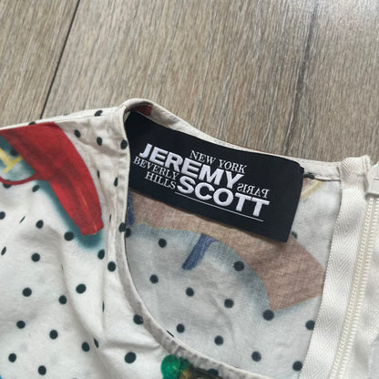Jeremy Scott SS2016 Watergun Dress is a white flared short dress featuring an all-over print of multicolored realistic waterguns in varying sizes and orientations. The design is set against a black dotted pattern layered across the fabric. It has a fitted upper section with a flared skirt silhouette and includes a black Jeremy Scott neck tag.