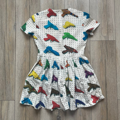 Jeremy Scott SS2016 Watergun Dress is a white flared short dress featuring an all-over print of multicolored realistic waterguns in varying sizes and orientations. The design is set against a black dotted pattern layered across the fabric. It has a fitted upper section with a flared skirt silhouette and includes a black Jeremy Scott neck tag.