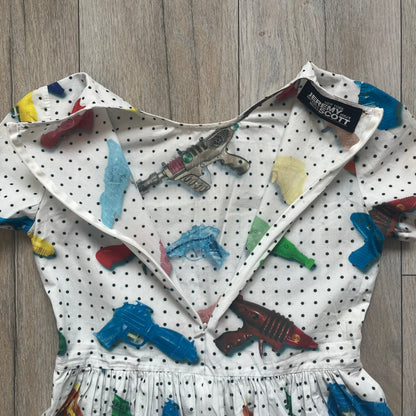 Jeremy Scott SS2016 Watergun Dress is a white flared short dress featuring an all-over print of multicolored realistic waterguns in varying sizes and orientations. The design is set against a black dotted pattern layered across the fabric. It has a fitted upper section with a flared skirt silhouette and includes a black Jeremy Scott neck tag.