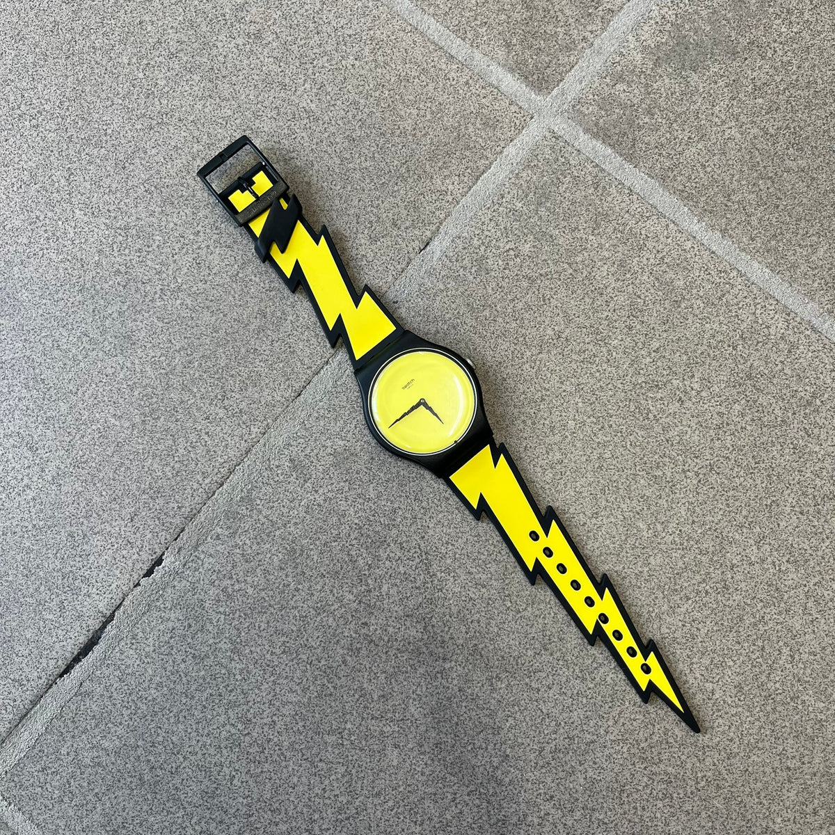 Jeremy Scott Swatch 2011 Lightning Flash Analog Watch (SUOZ104) is a limited-edition watch released in March 2011 from Jeremy Scott's first collaboration with Swatch. It features a black and yellow design with lightning bolt-shaped rubber straps, a yellow dial with a black case, lightning bolt-shaped hands, and a Jeremy Scott signature on the back.