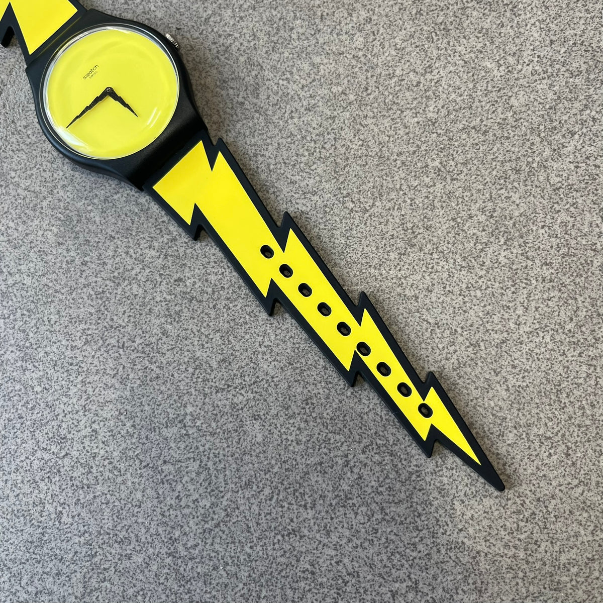 Jeremy Scott Swatch 2011 Lightning Flash Analog Watch (SUOZ104) is a limited-edition watch released in March 2011 from Jeremy Scott's first collaboration with Swatch. It features a black and yellow design with lightning bolt-shaped rubber straps, a yellow dial with a black case, lightning bolt-shaped hands, and a Jeremy Scott signature on the back.