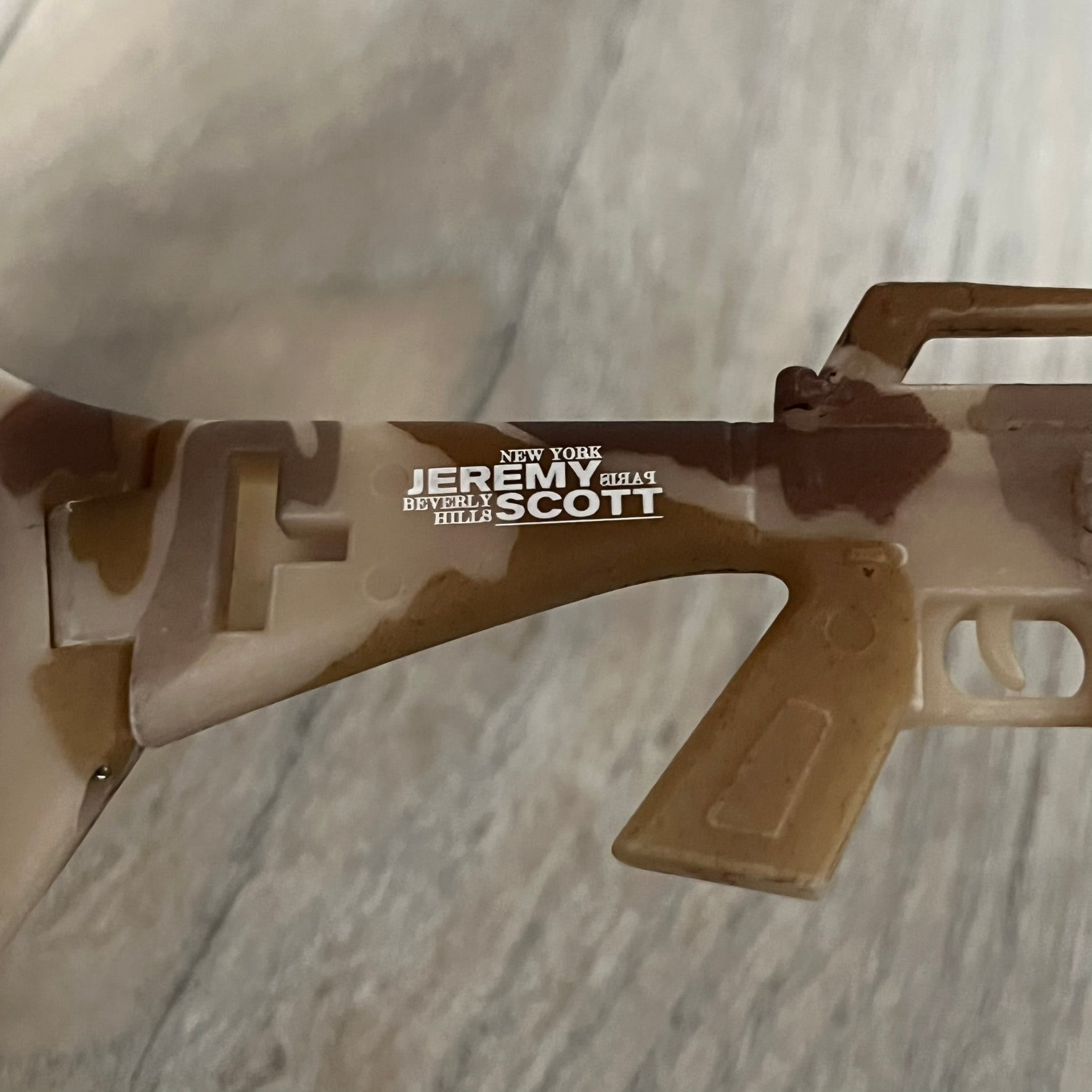 Jeremy Scott x Linda Farrow SS2013 Desert Camo M16 Rifle Sunglasses (JSARMYC4SUN) are a statement eyewear piece from the Spring/Summer 2013 collaboration. The design features full camo brown and beige frames with red/pink lenses, while the arms are constructed as detailed 3D M16 rifle shapes extending toward the front of the frame. The model includes Jeremy Scott branding on the inner arms and is produced in a high-detail acetate construction.