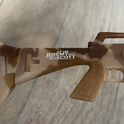 Jeremy Scott x Linda Farrow SS2013 Desert Camo M16 Rifle Sunglasses (JSARMYC4SUN) are a statement eyewear piece from the Spring/Summer 2013 collaboration. The design features full camo brown and beige frames with red/pink lenses, while the arms are constructed as detailed 3D M16 rifle shapes extending toward the front of the frame. The model includes Jeremy Scott branding on the inner arms and is produced in a high-detail acetate construction.