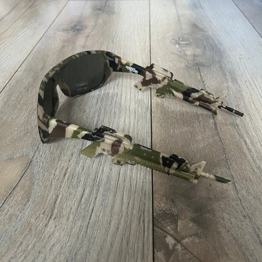 Jeremy Scott x Linda Farrow SS2013 Green Camo M16 Rifle Sunglasses (JSARMYC3SUN) are a statement eyewear piece from the Spring/Summer 2013 collaboration. The design features full camo green, brown, and beige frames with dark lenses, while the arms are constructed as detailed 3D M16 rifle shapes extending toward the front of the frame. The model includes Jeremy Scott branding on the inner arms and is produced in a high-detail acetate construction.