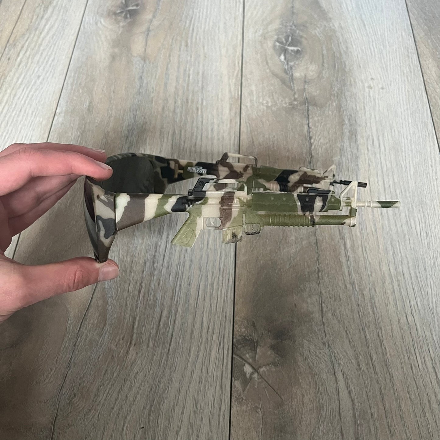 Jeremy Scott x Linda Farrow SS2013 Green Camo M16 Rifle Sunglasses (JSARMYC3SUN) are a statement eyewear piece from the Spring/Summer 2013 collaboration. The design features full camo green, brown, and beige frames with dark lenses, while the arms are constructed as detailed 3D M16 rifle shapes extending toward the front of the frame. The model includes Jeremy Scott branding on the inner arms and is produced in a high-detail acetate construction.