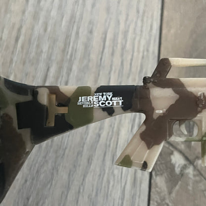 Jeremy Scott x Linda Farrow SS2013 Green Camo M16 Rifle Sunglasses (JSARMYC3SUN) are a statement eyewear piece from the Spring/Summer 2013 collaboration. The design features full camo green, brown, and beige frames with dark lenses, while the arms are constructed as detailed 3D M16 rifle shapes extending toward the front of the frame. The model includes Jeremy Scott branding on the inner arms and is produced in a high-detail acetate construction.