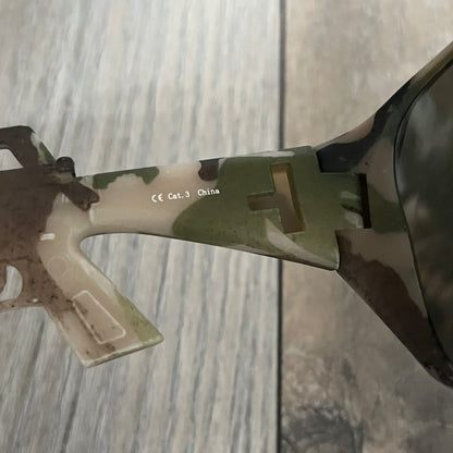 Jeremy Scott x Linda Farrow SS2013 Green Camo M16 Rifle Sunglasses (JSARMYC3SUN) are a statement eyewear piece from the Spring/Summer 2013 collaboration. The design features full camo green, brown, and beige frames with dark lenses, while the arms are constructed as detailed 3D M16 rifle shapes extending toward the front of the frame. The model includes Jeremy Scott branding on the inner arms and is produced in a high-detail acetate construction.