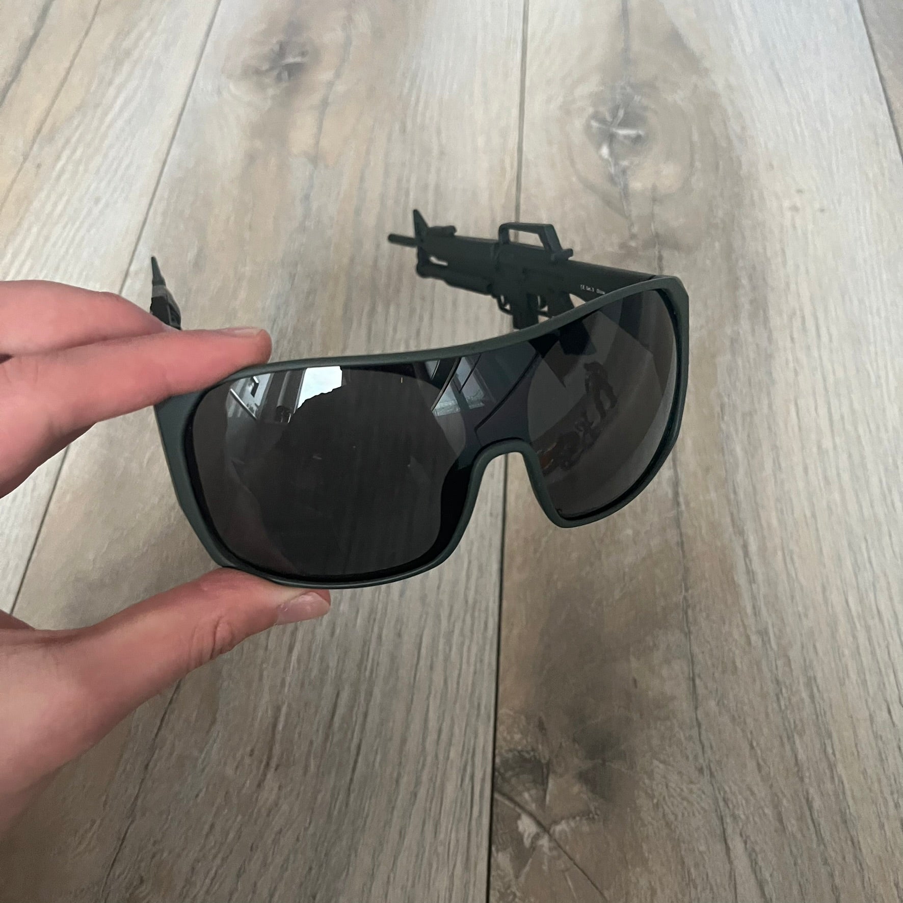 Jeremy Scott x Linda Farrow SS2013 Matt Khaki M16 Rifle Sunglasses (JSARMYC5SUN) are a statement eyewear piece from the Spring/Summer 2013 collaboration. The design features full matt dark khaki frames with black lenses, while the arms are constructed as detailed 3D M16 rifle shapes extending toward the front of the frame. The model includes Jeremy Scott branding on the inner arms and is produced in a high-detail acetate construction.