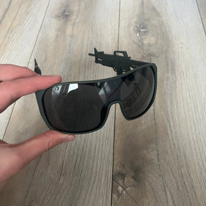 Jeremy Scott x Linda Farrow SS2013 Matt Khaki M16 Rifle Sunglasses (JSARMYC5SUN) are a statement eyewear piece from the Spring/Summer 2013 collaboration. The design features full matt dark khaki frames with black lenses, while the arms are constructed as detailed 3D M16 rifle shapes extending toward the front of the frame. The model includes Jeremy Scott branding on the inner arms and is produced in a high-detail acetate construction.