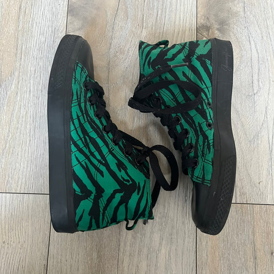 Jeremy Scott x adidas 2014 Nizza Hi “Forest Green” Sneakers are made with a forest green canvas upper featuring an all-over black tiger print. The high-top shoes include rubber soles and standard adidas Nizza tooling. This model was released in 2014 in a green and black colorway as part of the Jeremy Scott x adidas collaboration.