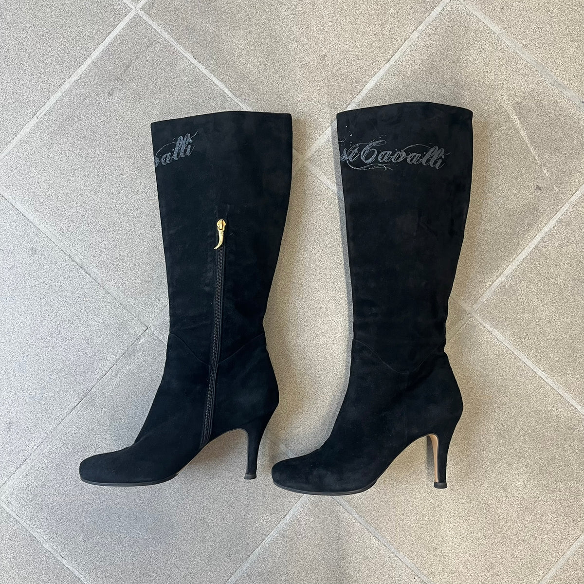 Just Cavalli 2000's Black Suede Boots are made in Italy, featuring black suede uppers with a beige sole and "Just Cavalli" branding on the bottom. A glittery silver "Just Cavalli" text logo appears at the top of the boot, with a side zipper running from the midpoint down, finished with a gold Just Cavalli horn zipper pull.