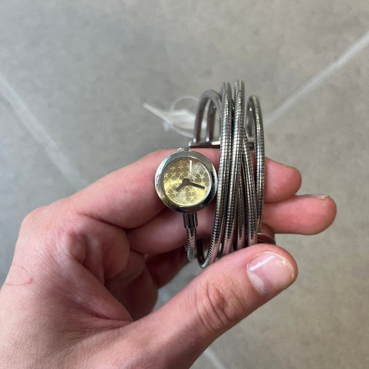 Just Cavalli 2000’s Yellow Stars Bracelet Watch is a stretch metal bracelet watch from Just Cavalli featuring multiple intersecting silver-tone coil bands that create a layered wrap-around design. One end of the bracelet holds a small round analog watch with a yellow dial finished with a textured star pattern, while the opposite end features a circular metal element engraved with the JC logo. The bracelet uses a spring-style stretch construction instead of a clasp, allowing it to expand to fit the wrist. Th