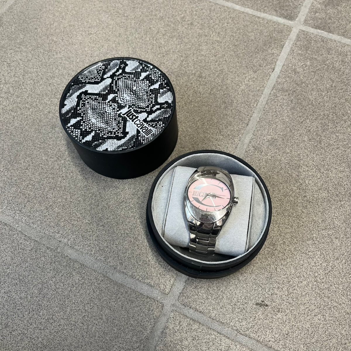 Just Cavalli 2010’s Pink Dial Analog Watch is a stainless steel watch with a raised oval case and an oval pink dial with a large silver Just Cavalli logo. The clasp is Just Cavalli branded, and the watch comes with its box and papers.