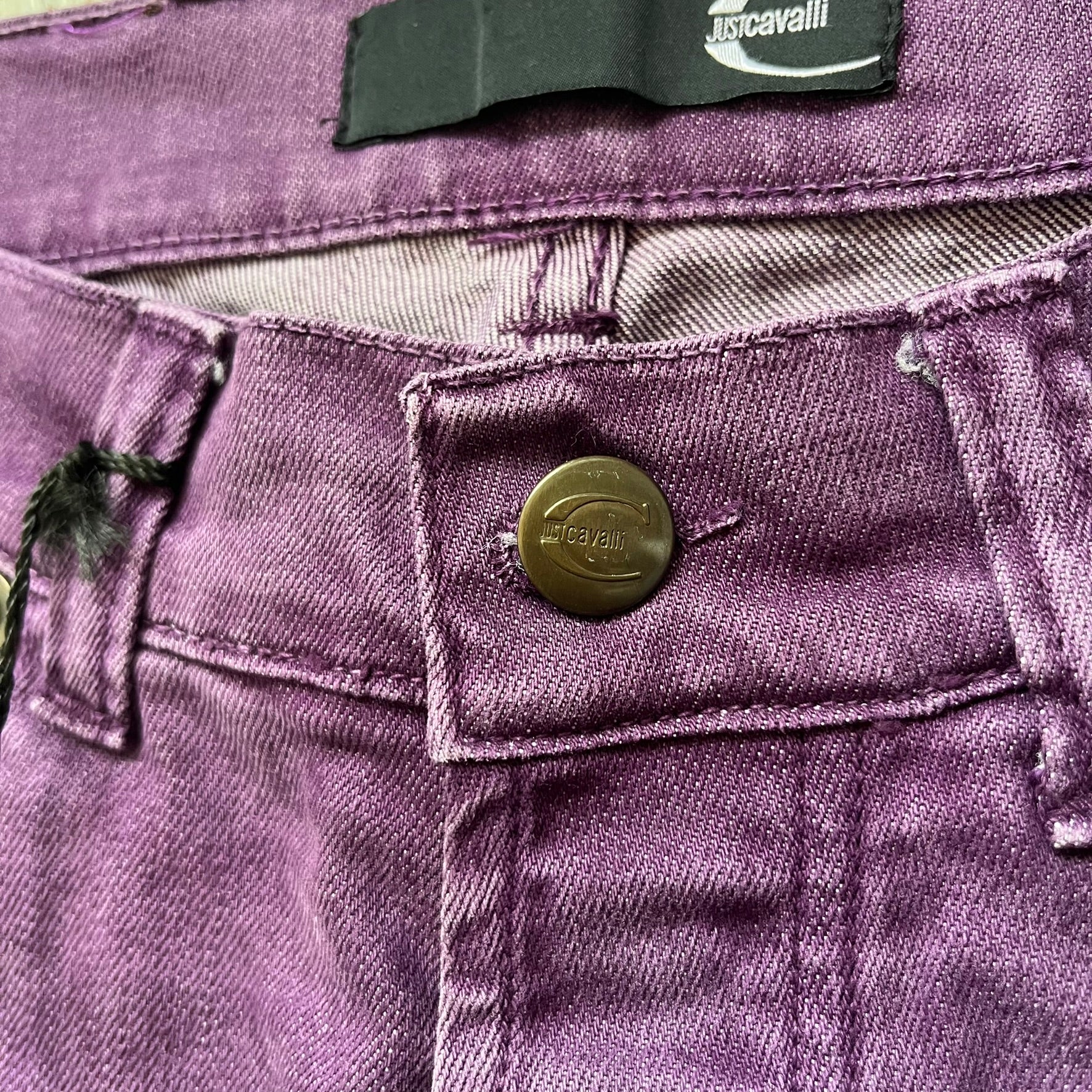 Just Cavalli 2010s Purple Slim Jeans are slim-fit denim jeans finished in a full purple colorway. The pair features classic front and back pocket construction, branded metal button closure, and small metal stud details on the pockets. Additional details include a rear leather Just Cavalli logo patch, interior Just Cavalli tag, and original paper tag.