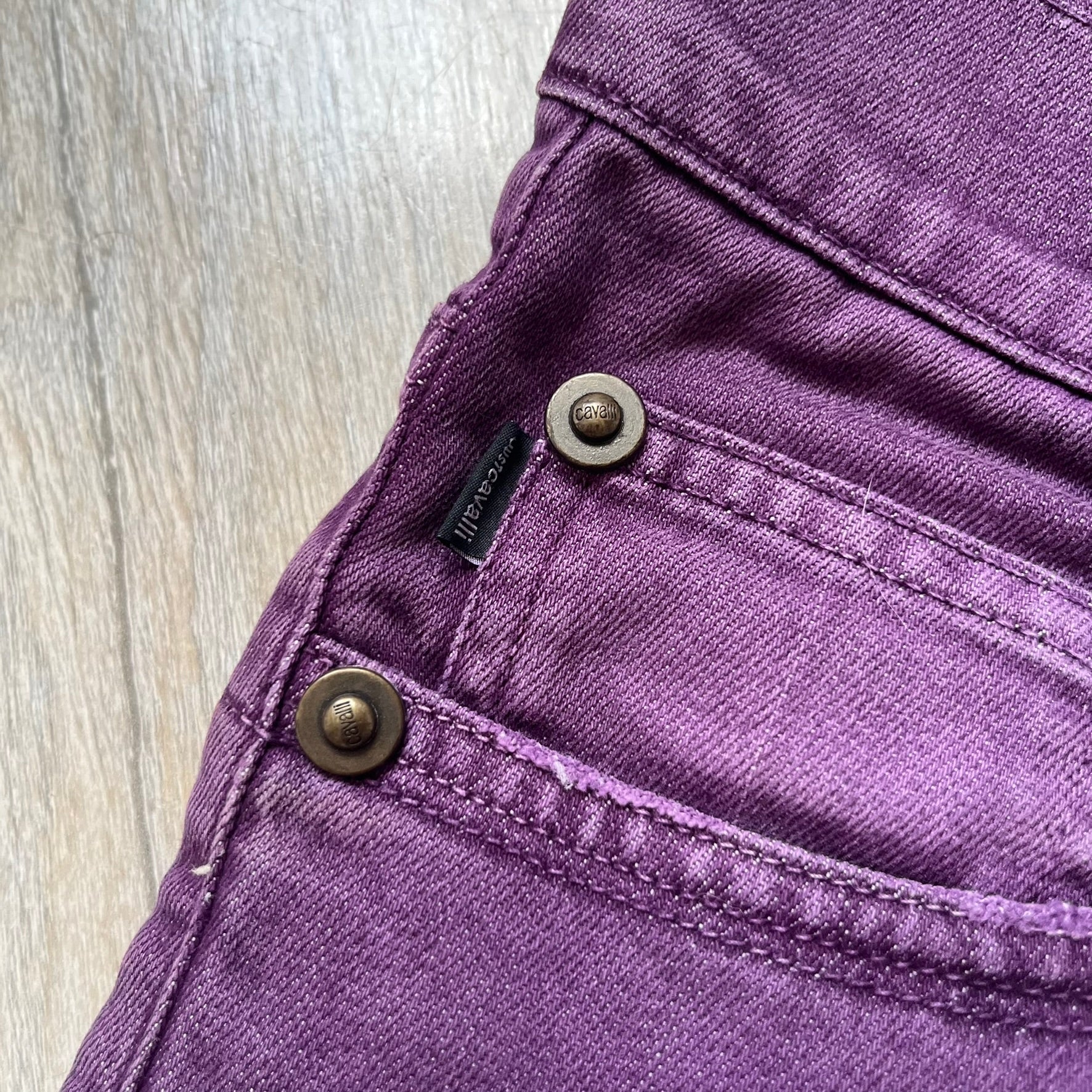 Just Cavalli 2010s Purple Slim Jeans are slim-fit denim jeans finished in a full purple colorway. The pair features classic front and back pocket construction, branded metal button closure, and small metal stud details on the pockets. Additional details include a rear leather Just Cavalli logo patch, interior Just Cavalli tag, and original paper tag.