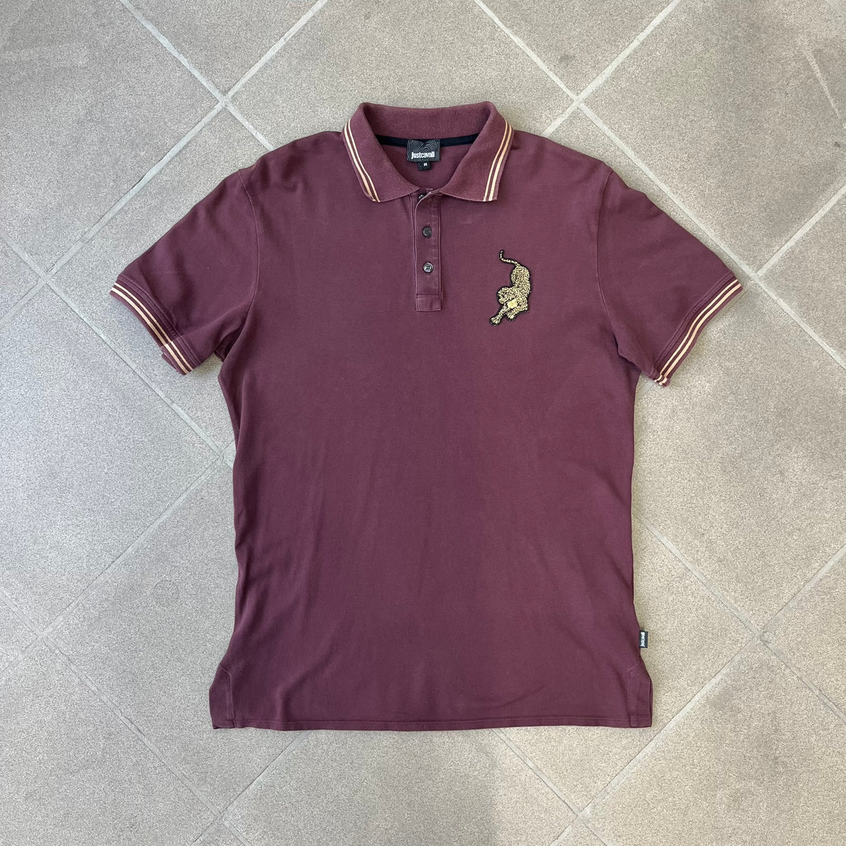 Just Cavalli 2017 Leopard Patch Polo is a burgundy, 100% cotton polo with gold/yellow trim along the hems and collar, featuring a roaring leopard patch on the chest and a small Cavalli side tag. It has side vents at the hem, typical of the polo style.