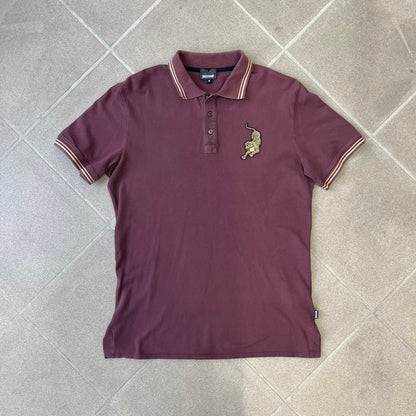 Just Cavalli 2017 Leopard Patch Polo is a burgundy, 100% cotton polo with gold/yellow trim along the hems and collar, featuring a roaring leopard patch on the chest and a small Cavalli side tag. It has side vents at the hem, typical of the polo style.