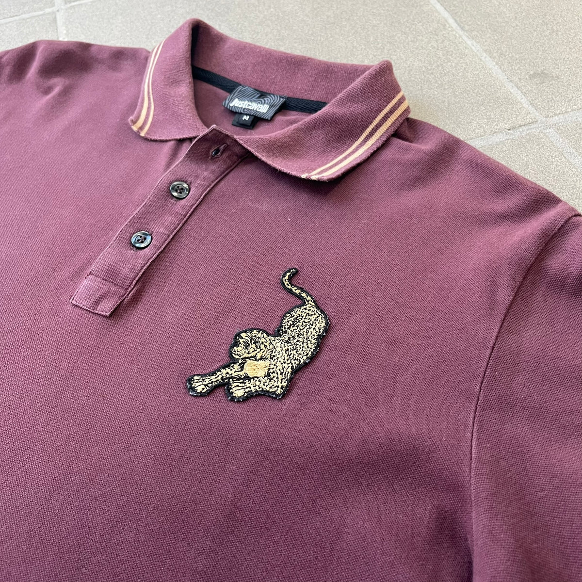 Just Cavalli 2017 Leopard Patch Polo is a burgundy, 100% cotton polo with gold/yellow trim along the hems and collar, featuring a roaring leopard patch on the chest and a small Cavalli side tag. It has side vents at the hem, typical of the polo style.