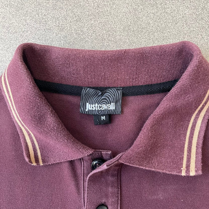 Just Cavalli 2017 Leopard Patch Polo is a burgundy, 100% cotton polo with gold/yellow trim along the hems and collar, featuring a roaring leopard patch on the chest and a small Cavalli side tag. It has side vents at the hem, typical of the polo style.