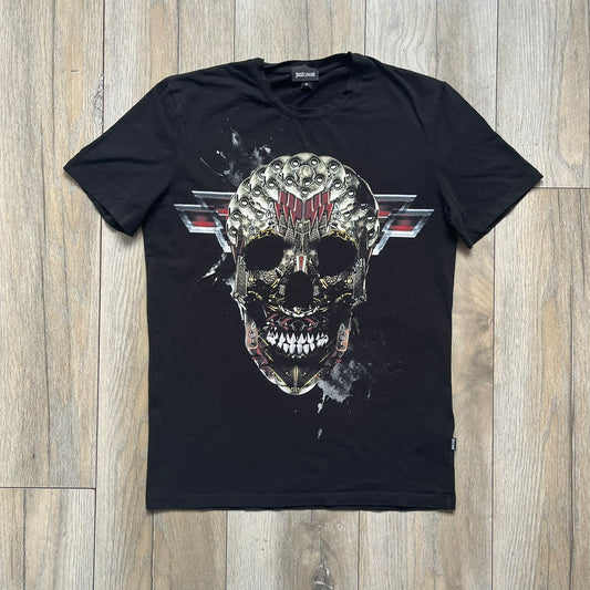 Just Cavalli AW2016 Metal Skull Print Black Tee is a black cotton T-shirt featuring a detailed front graphic of a metallic-style skull composed of gold, silver, and red plate-like elements. The design includes red accents and white splatter effects in the background for contrast. It comes with a black Just Cavalli neck tag and a small side tag.