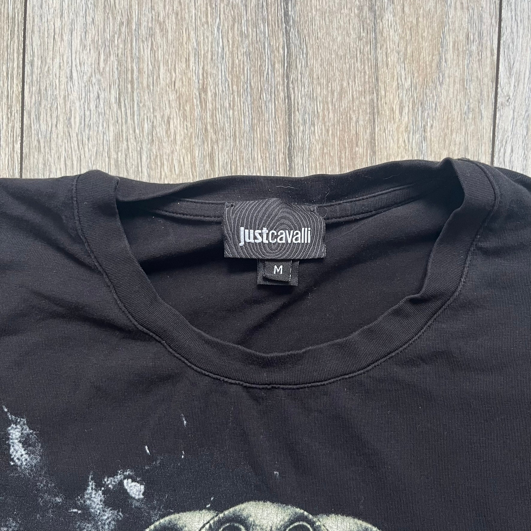 Just Cavalli AW2016 Metal Skull Print Black Tee is a black cotton T-shirt featuring a detailed front graphic of a metallic-style skull composed of gold, silver, and red plate-like elements. The design includes red accents and white splatter effects in the background for contrast. It comes with a black Just Cavalli neck tag and a small side tag.