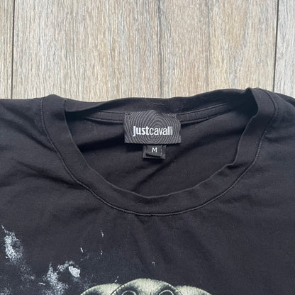 Just Cavalli AW2016 Metal Skull Print Black Tee is a black cotton T-shirt featuring a detailed front graphic of a metallic-style skull composed of gold, silver, and red plate-like elements. The design includes red accents and white splatter effects in the background for contrast. It comes with a black Just Cavalli neck tag and a small side tag.
