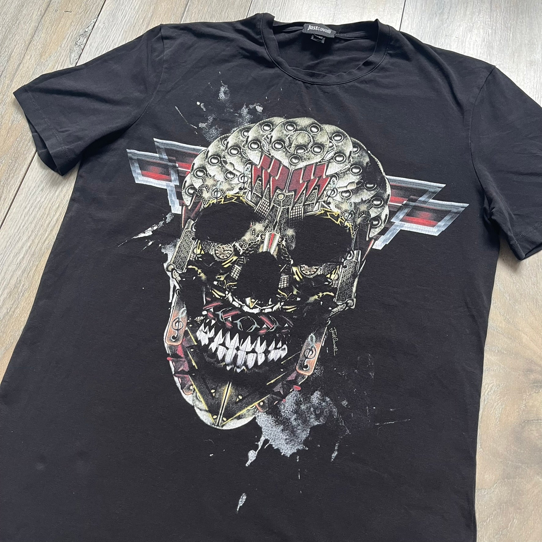 Just Cavalli AW2016 Metal Skull Print Black Tee is a black cotton T-shirt featuring a detailed front graphic of a metallic-style skull composed of gold, silver, and red plate-like elements. The design includes red accents and white splatter effects in the background for contrast. It comes with a black Just Cavalli neck tag and a small side tag.