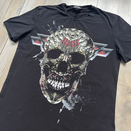 Just Cavalli AW2016 Metal Skull Print Black Tee is a black cotton T-shirt featuring a detailed front graphic of a metallic-style skull composed of gold, silver, and red plate-like elements. The design includes red accents and white splatter effects in the background for contrast. It comes with a black Just Cavalli neck tag and a small side tag.