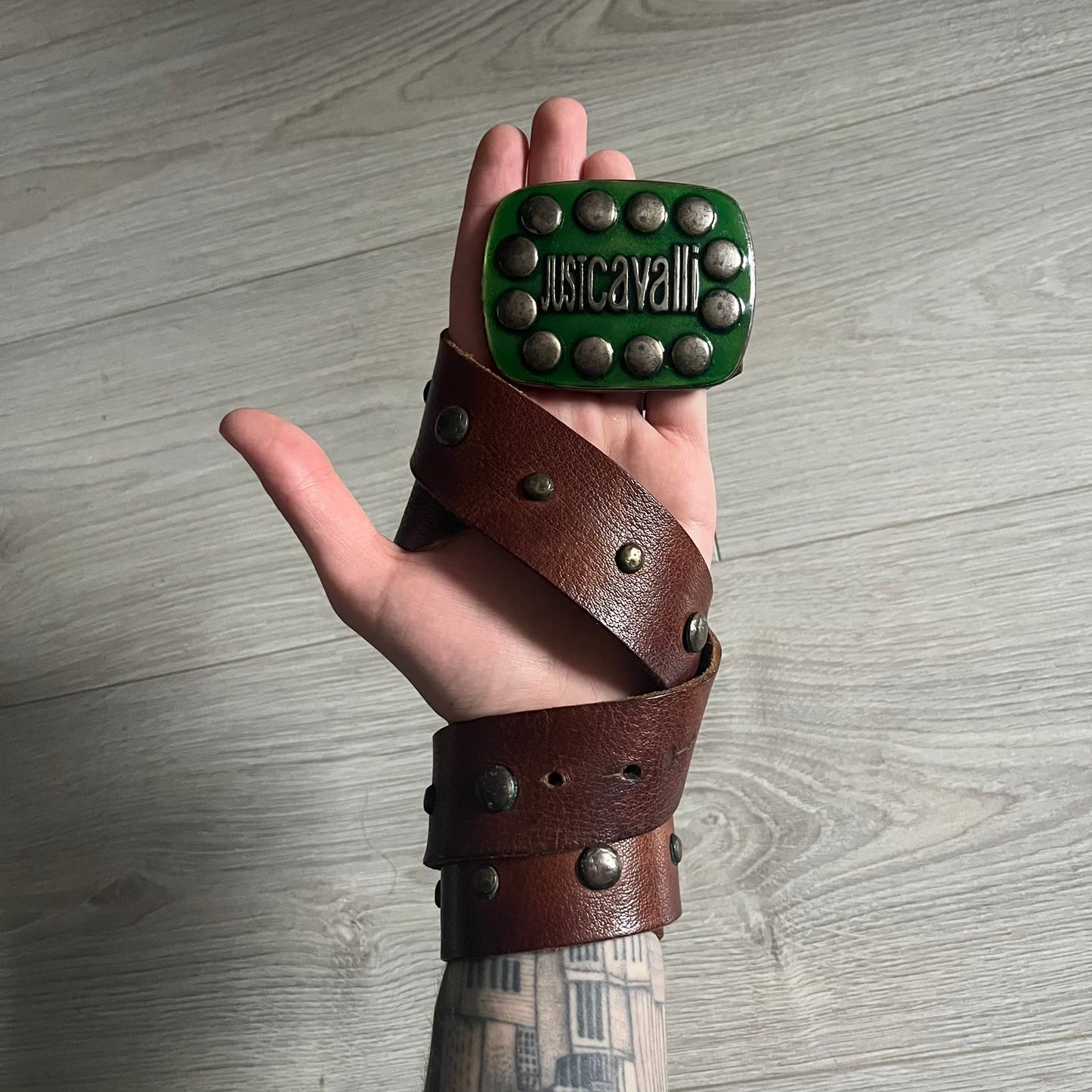 Just Cavalli Green Metal Buckle Studded Belt featuring a shiny green metallic buckle with the Just Cavalli logo in the center, surrounded by metallic studs. The brown leather strap is accented with a line of metallic studs along its length.