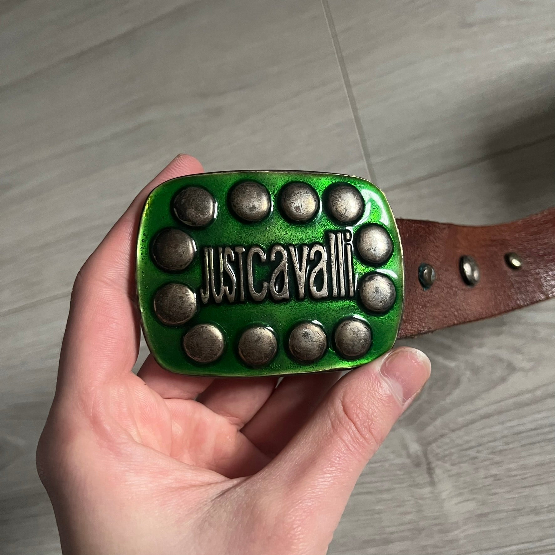Just Cavalli Green Metal Buckle Studded Belt featuring a shiny green metallic buckle with the Just Cavalli logo in the center, surrounded by metallic studs. The brown leather strap is accented with a line of metallic studs along its length.