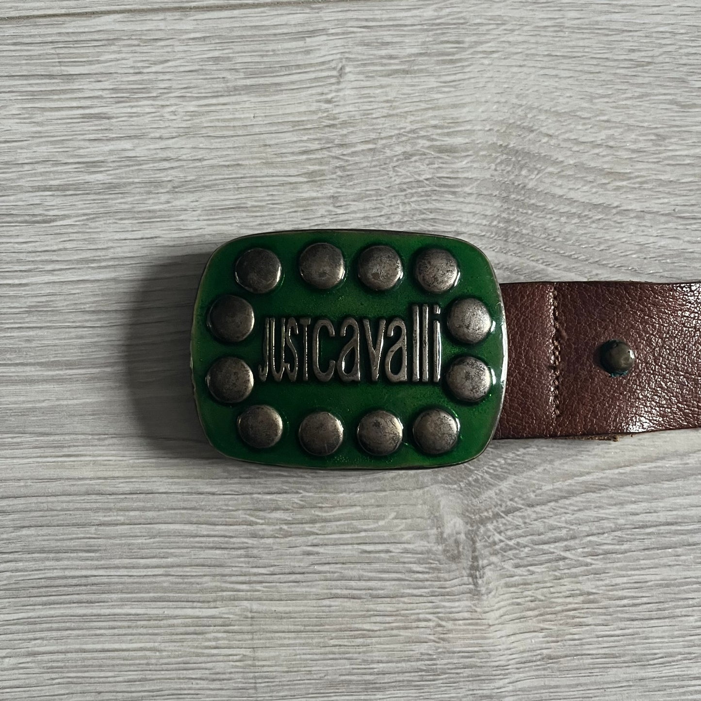 Just Cavalli Green Metal Buckle Studded Belt featuring a shiny green metallic buckle with the Just Cavalli logo in the center, surrounded by metallic studs. The brown leather strap is accented with a line of metallic studs along its length.