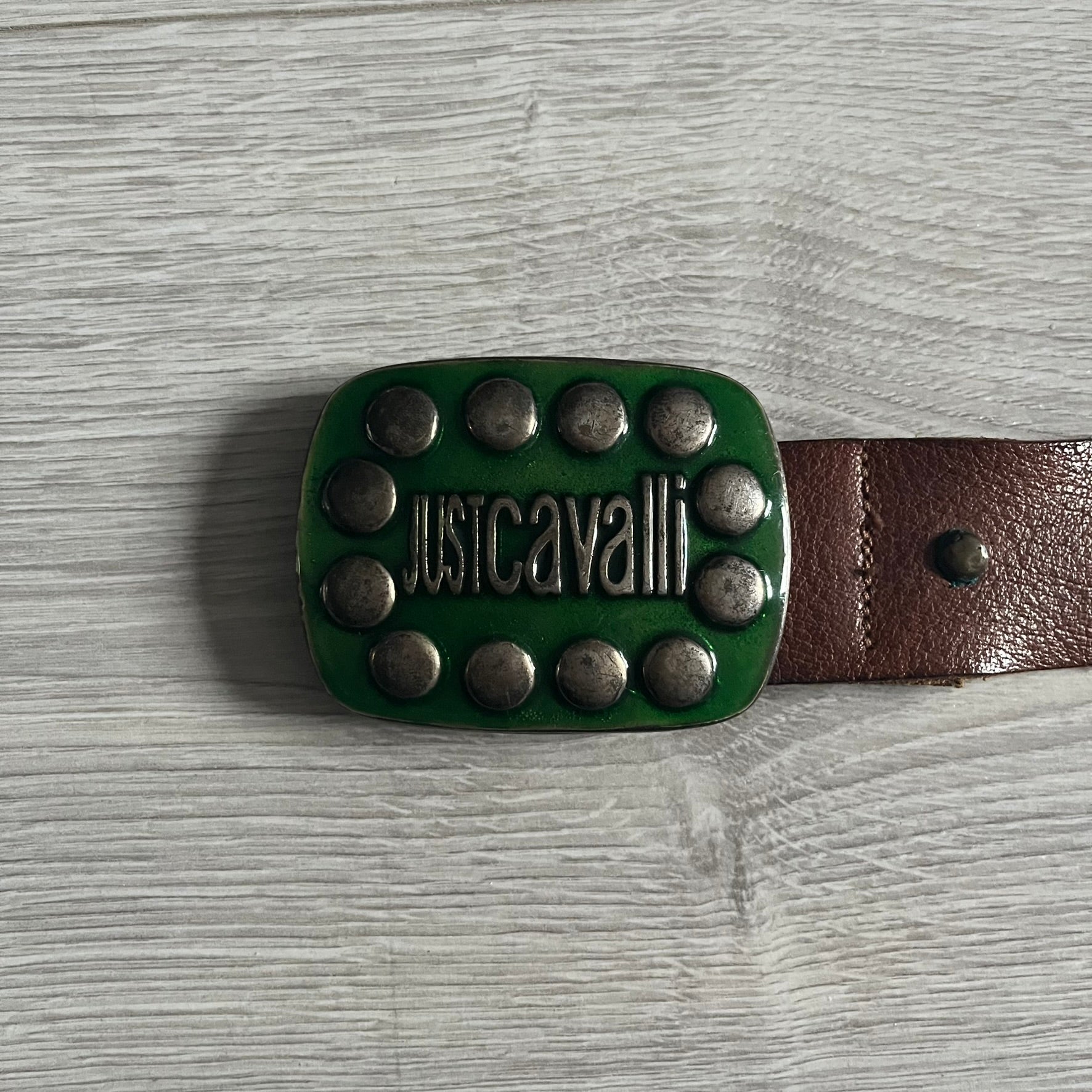 Just Cavalli Green Metal Buckle Studded Belt featuring a shiny green metallic buckle with the Just Cavalli logo in the center, surrounded by metallic studs. The brown leather strap is accented with a line of metallic studs along its length.