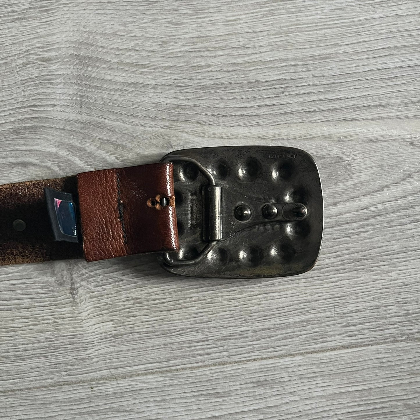 Just Cavalli Green Metal Buckle Studded Belt featuring a shiny green metallic buckle with the Just Cavalli logo in the center, surrounded by metallic studs. The brown leather strap is accented with a line of metallic studs along its length.