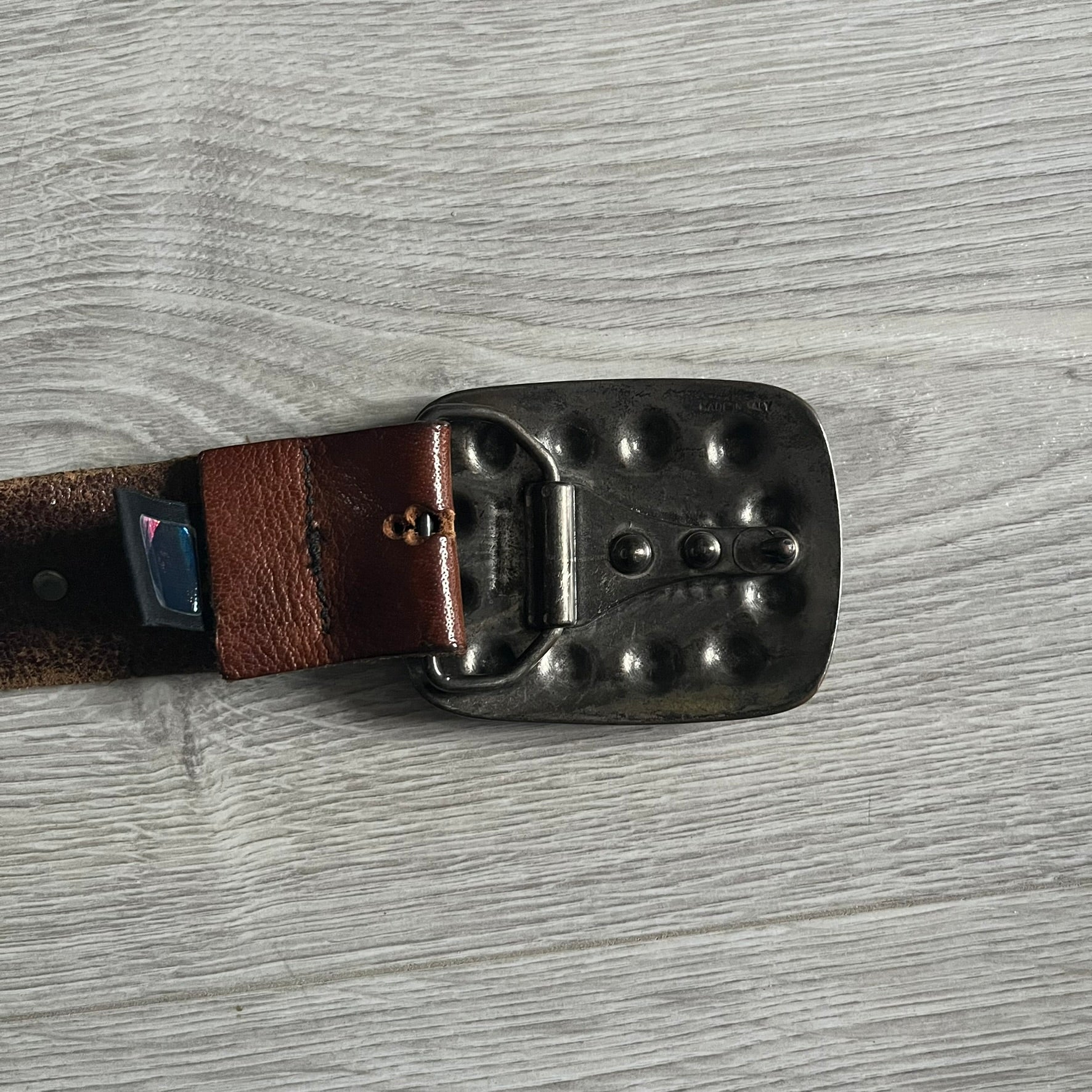 Just Cavalli Green Metal Buckle Studded Belt featuring a shiny green metallic buckle with the Just Cavalli logo in the center, surrounded by metallic studs. The brown leather strap is accented with a line of metallic studs along its length.