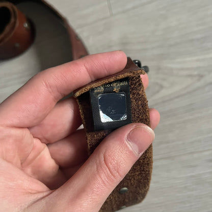 Just Cavalli Green Metal Buckle Studded Belt featuring a shiny green metallic buckle with the Just Cavalli logo in the center, surrounded by metallic studs. The brown leather strap is accented with a line of metallic studs along its length.