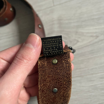 Just Cavalli Green Metal Buckle Studded Belt featuring a shiny green metallic buckle with the Just Cavalli logo in the center, surrounded by metallic studs. The brown leather strap is accented with a line of metallic studs along its length.