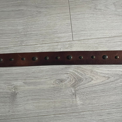 Just Cavalli Green Metal Buckle Studded Belt featuring a shiny green metallic buckle with the Just Cavalli logo in the center, surrounded by metallic studs. The brown leather strap is accented with a line of metallic studs along its length.