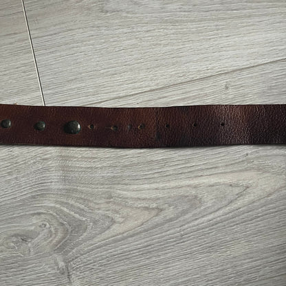 Just Cavalli Green Metal Buckle Studded Belt featuring a shiny green metallic buckle with the Just Cavalli logo in the center, surrounded by metallic studs. The brown leather strap is accented with a line of metallic studs along its length.