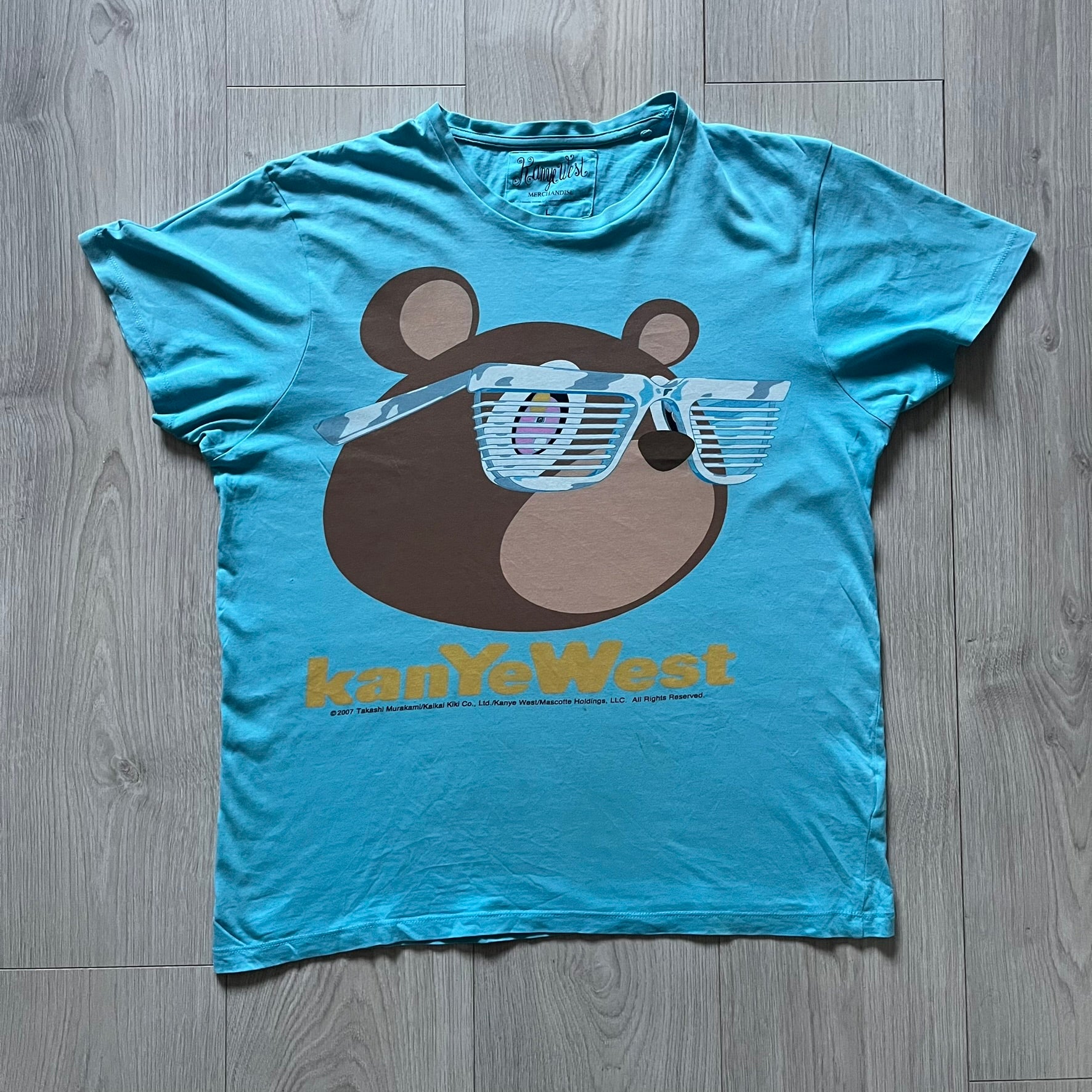 Kanye West 2012 Takashi Murakami 'Glow In The Dark Tour' Tee in light blue features the iconic bear with shutter glasses on the front. Below the graphic is yellow "kanYeWest" text and the 2007 Takashi Murakami/Kanye West licensing. Official merchandise from the 2012, this collectible tee includes the neck tag with Kanye logo and Mascotte Holdings licensing.