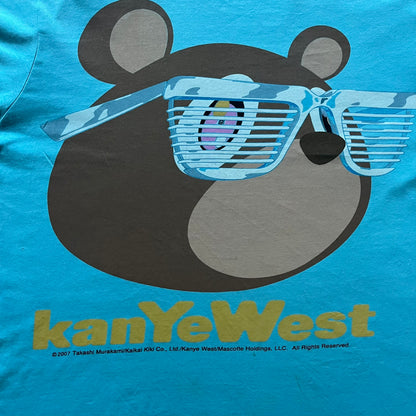 Kanye West 2012 Takashi Murakami 'Glow In The Dark Tour' Tee in light blue features the iconic bear with shutter glasses on the front. Below the graphic is yellow "kanYeWest" text and the 2007 Takashi Murakami/Kanye West licensing. Official merchandise from the 2012, this collectible tee includes the neck tag with Kanye logo and Mascotte Holdings licensing.