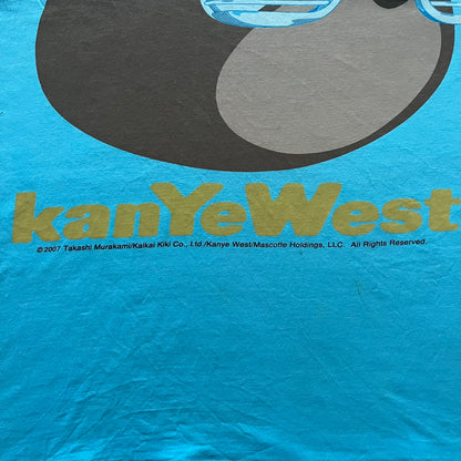 Kanye West 2012 Takashi Murakami 'Glow In The Dark Tour' Tee in light blue features the iconic bear with shutter glasses on the front. Below the graphic is yellow "kanYeWest" text and the 2007 Takashi Murakami/Kanye West licensing. Official merchandise from the 2012, this collectible tee includes the neck tag with Kanye logo and Mascotte Holdings licensing.