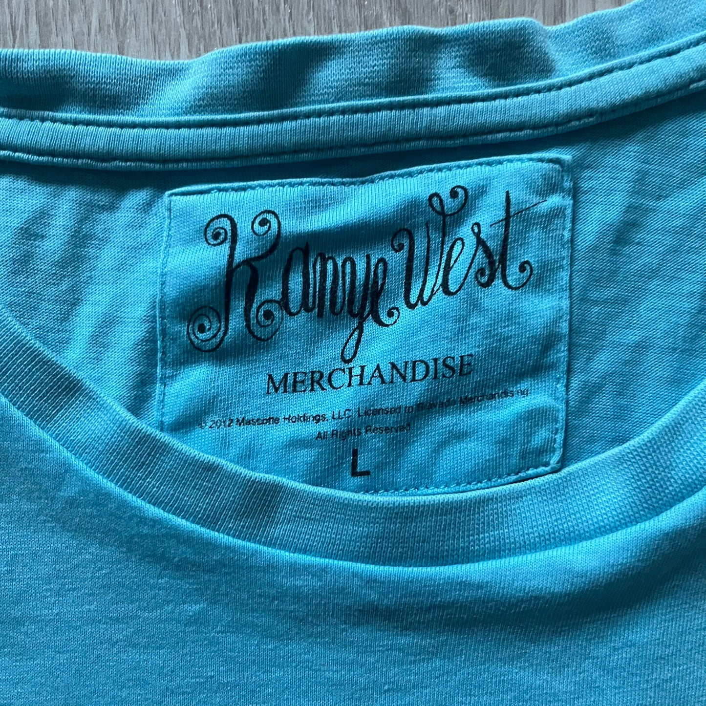 Kanye West 2012 Takashi Murakami 'Glow In The Dark Tour' Tee in light blue features the iconic bear with shutter glasses on the front. Below the graphic is yellow "kanYeWest" text and the 2007 Takashi Murakami/Kanye West licensing. Official merchandise from the 2012, this collectible tee includes the neck tag with Kanye logo and Mascotte Holdings licensing.